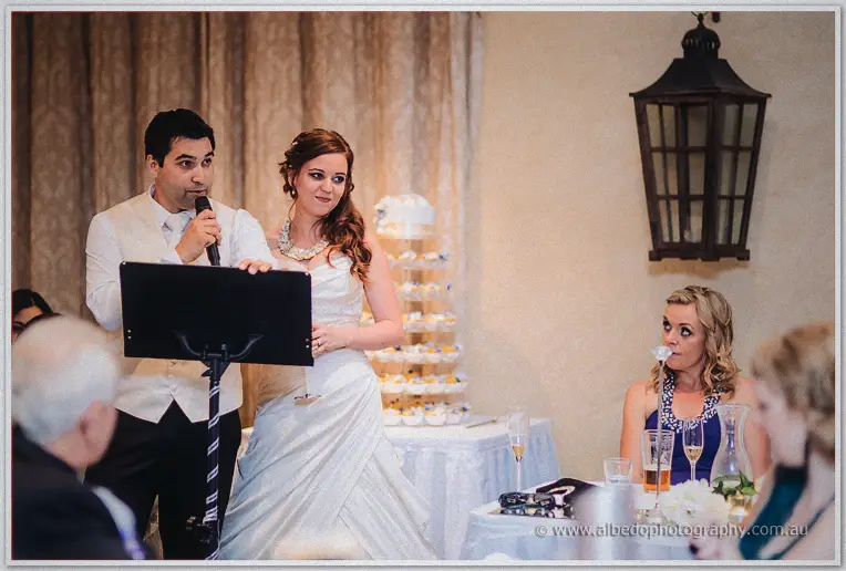 Craig speaks at the lectern with Amanda beside him and the tiered cupcake tower glowing behind