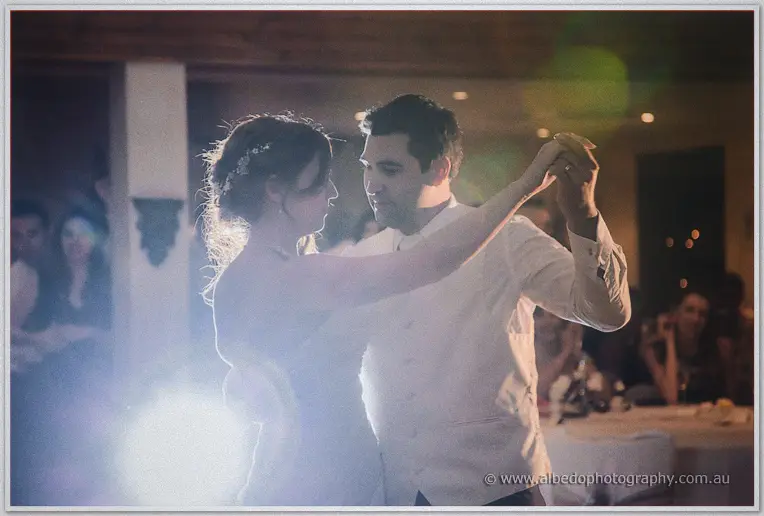 Bride and groom forehead to forehead during a spotlight first dance, wedding guests blurred behind them