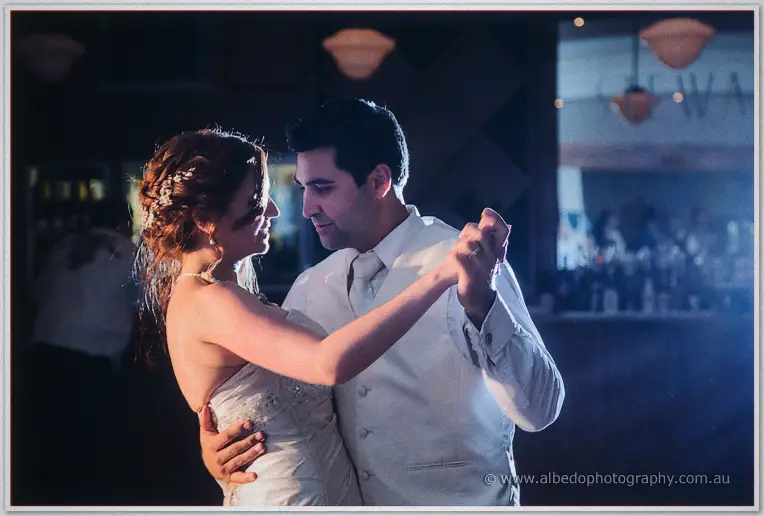 Amanda and Craig dance cheek to cheek in soft evening light with the reception bar glowing behind