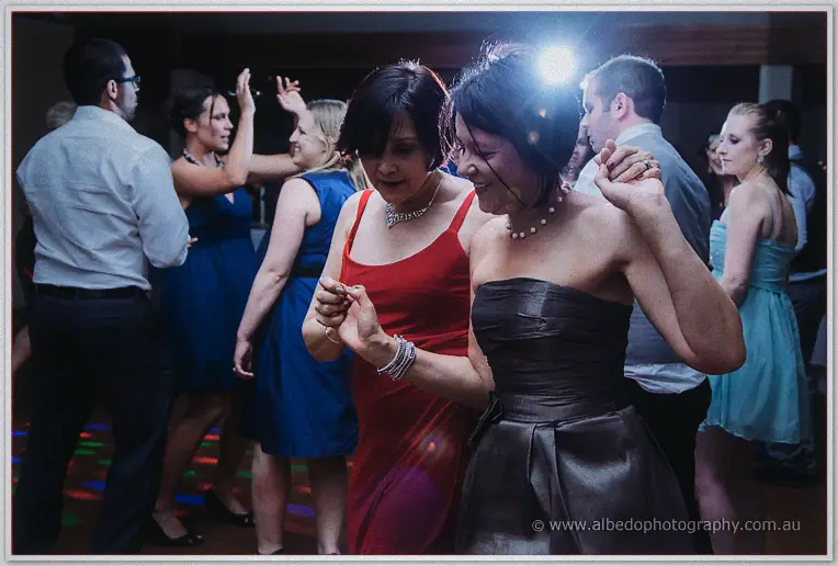 Two female guests in red and charcoal dresses dance together in the flash-lit wedding crowd