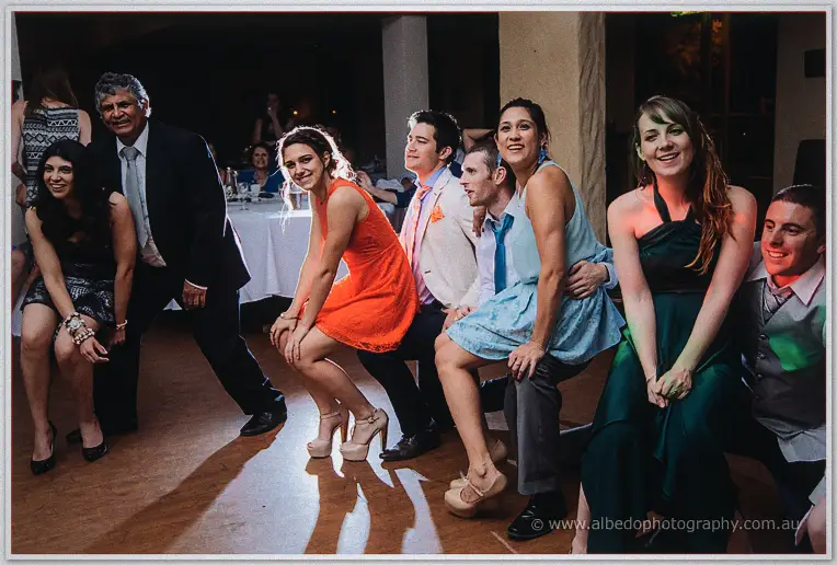 Row of wedding guests crouch shoulder to shoulder on the dance floor striking a playful pose