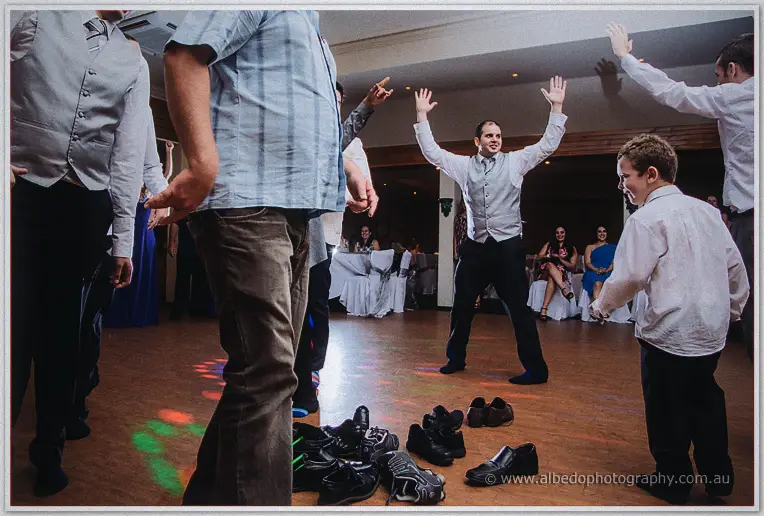 A groomsman throws both arms up dancing beside a young boy, shoes heaped on the floor before them