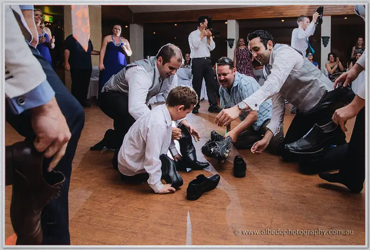Groomsmen kneel to sort a pile of shoes with the page boy while guests cheer from the sidelines