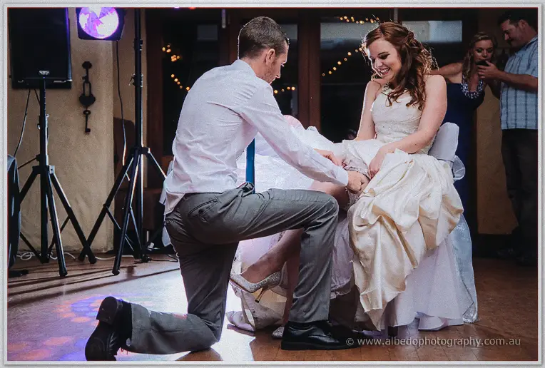 Garter toss moment as a groomsman kneels to remove the garter while the seated bride laughs