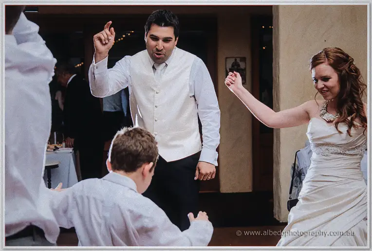 Groom in white vest dances with a young page boy while Amanda laughs beside them at the reception