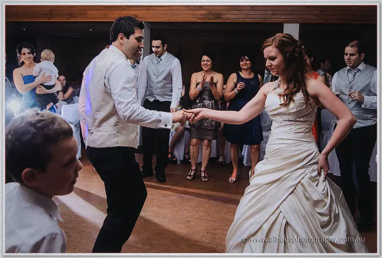 Amanda and Craig dance hand in hand surrounded by clapping guests at the evening reception