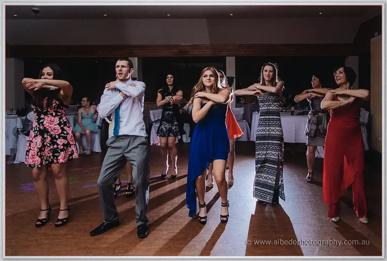 Wedding guests in blue, red and floral dresses dance in a line with arms crossed on the floor
