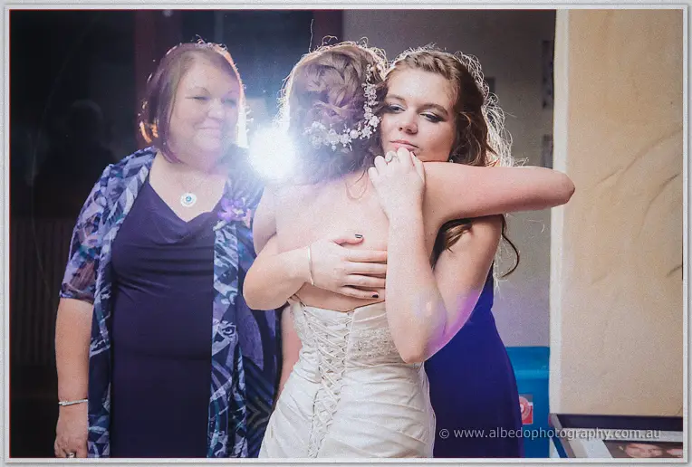 Bride hugs a young bridesmaid close as her mother looks on warmly at the reception
