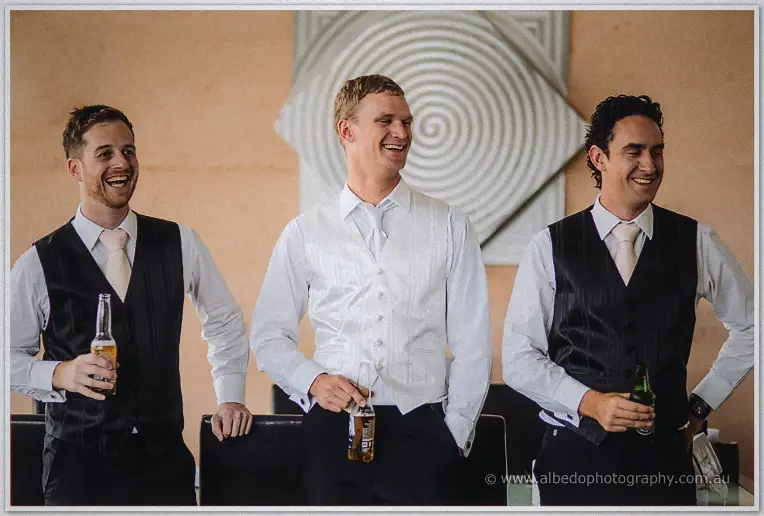 The groom flanked by two laughing groomsmen in black vests, beers in hand, by a spiral wall art piece
