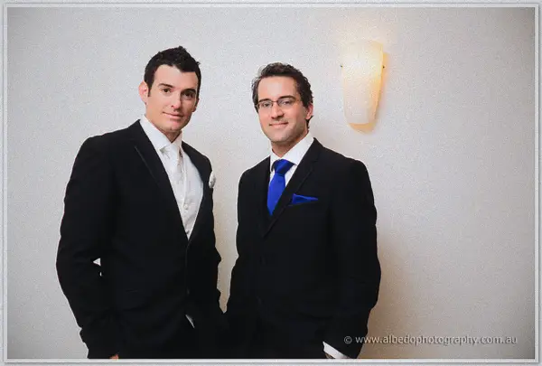 Joe and his best man in dark suits with blue ties pose side by side against a white wall