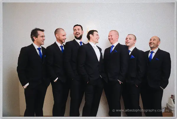 Joe laughs with his groomsmen, all in navy suits and blue ties, lined up by a white wall