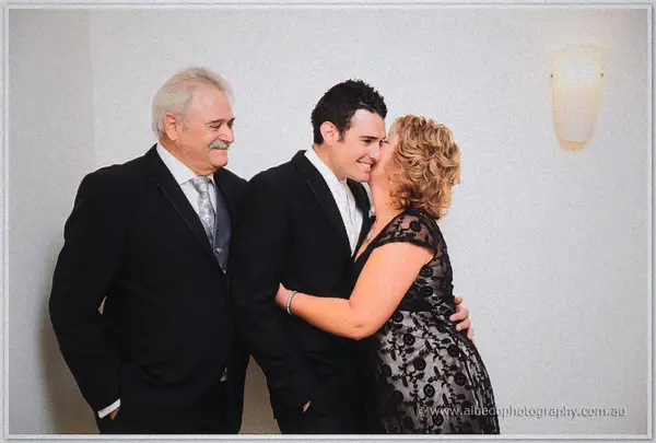 Joe's parents flank him by a wall lamp as his mother kisses his cheek before the ceremony