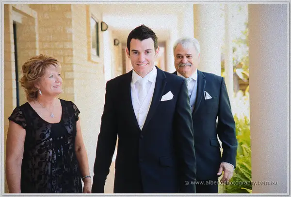Groom Joe walks a sunlit veranda with his smiling parents on either side
