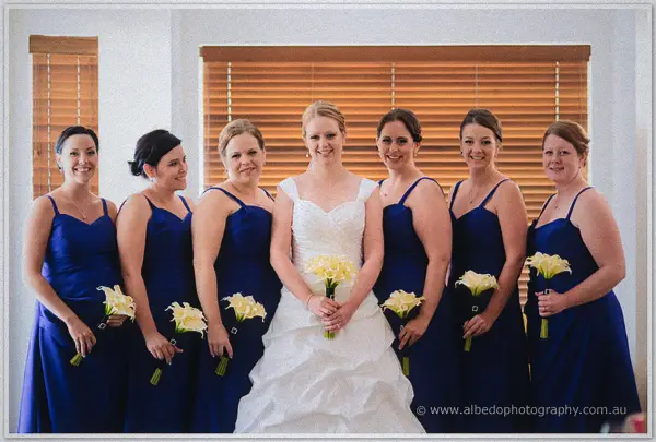 Cayla and her six bridesmaids in blue dresses hold cream calla lilies by a timber blind