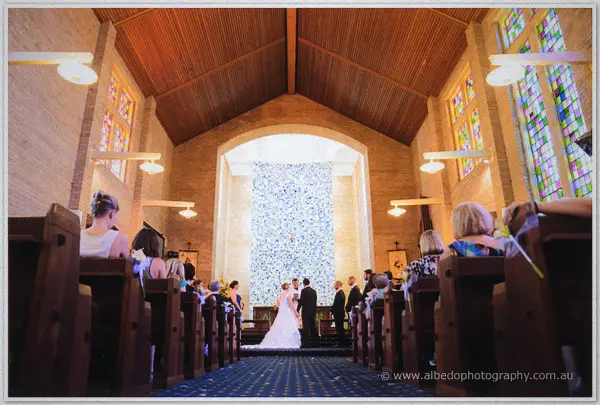 Cayla and Joe hold hands at the altar as guests watch beneath the cathedral's bright reredos