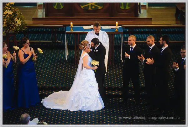Joe kisses Cayla at the altar while groomsmen applaud and bridesmaids in blue cheer