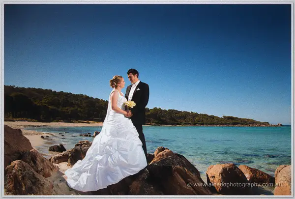 Cayla and Joe stand on granite rocks above a turquoise bay under a deep blue sky