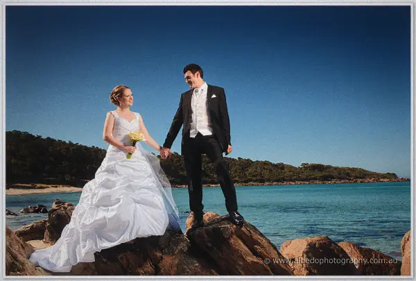 The newlyweds hold hands on coastal rocks, her train pooling beside the clear water
