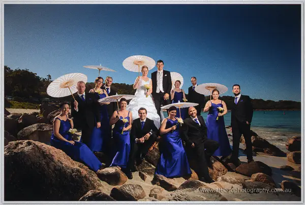Bridal party with paper parasols clusters on beach rocks around the laughing couple