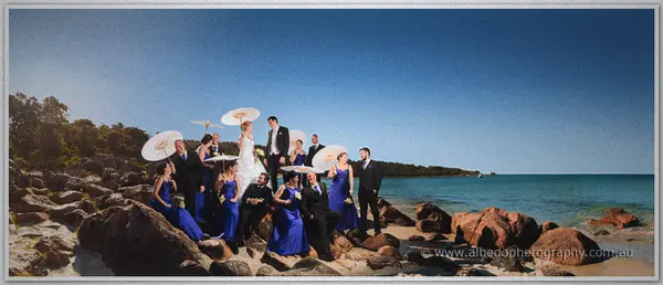 Panoramic bridal party photo with parasols on granite boulders beside the bay