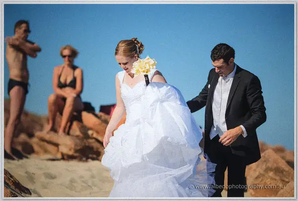 Joe steadies Cayla's billowing gown on the sand as bemused beachgoers watch
