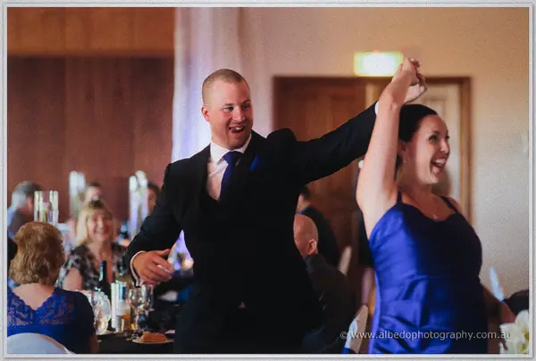 A groomsman in black and a woman in royal blue dance with arms raised among laughing guests at the reception