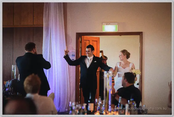 Joe strides through the doorway with an arm raised as Cayla follows with her cream bouquet, guests watching