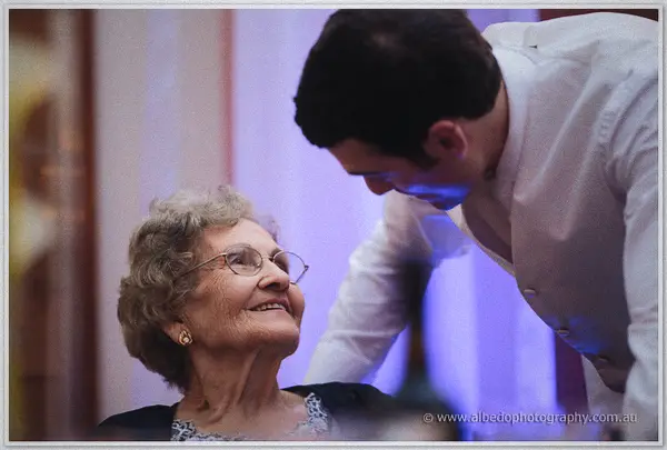 An elderly guest in glasses laughs with the groom in his white shirt under warm reception lights