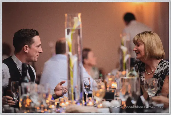 The groom chats with a guest at a candlelit table beneath tall calla lily centerpieces