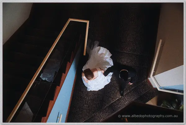 Seen from the staircase above, Cayla and Joe stand close together on a patterned landing in dramatic light