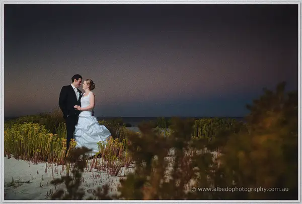 Cayla and Joe embrace among coastal shrubs on the dunes above a calm sea under a purple twilight sky