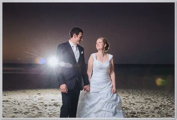 Laughing hand in hand on the night beach, the couple is rimmed by a bright flare of backlight between them