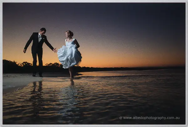 Joe leads Cayla by the hand as she gathers her gown and wades barefoot through shallow sunset-lit shallows