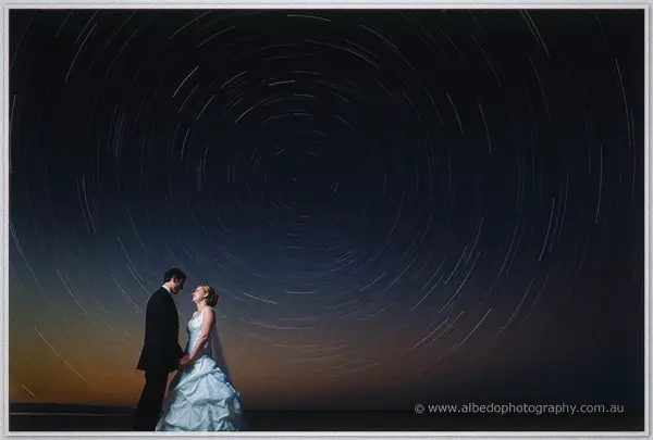 Cayla and Joe hold hands beneath sweeping circular star trails over a glowing dusk horizon