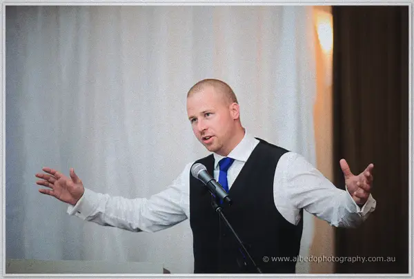 A groomsman gestures expressively mid-speech at the microphone before a curtain of white drapery
