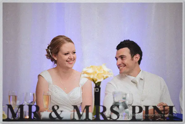 Cayla and Joe laugh at their bridal table behind a Mr and Mrs sign, yellow callas and champagne flutes nearby