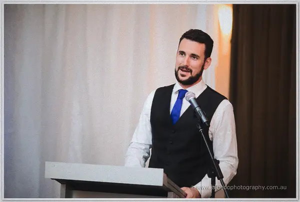 A bearded groomsman in vest and blue tie delivers his speech from the lectern against the curtained wall