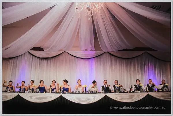 The full bridal party raises glasses along the draped head table beneath the chandelier and purple uplighting