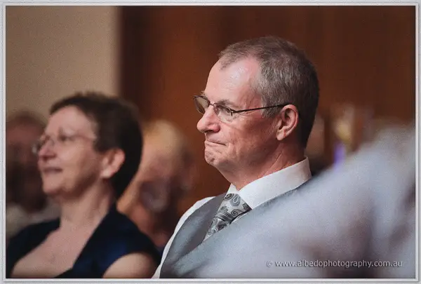 A grey-haired guest in glasses watches the speeches with quiet emotion beside a smiling companion