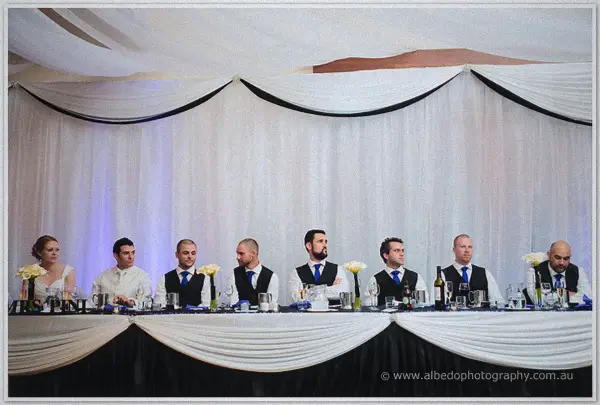The bridal party lines the head table in black vests, blue ties and yellow roses beneath the white draping