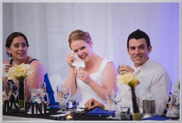 Cayla dabs happy tears between a laughing bridesmaid and a grinning Joe at the head table