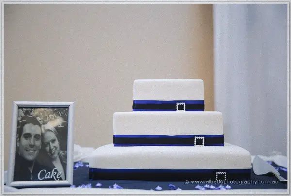 A three-tier white cake with navy ribbon and buckles stands beside a framed portrait of the couple