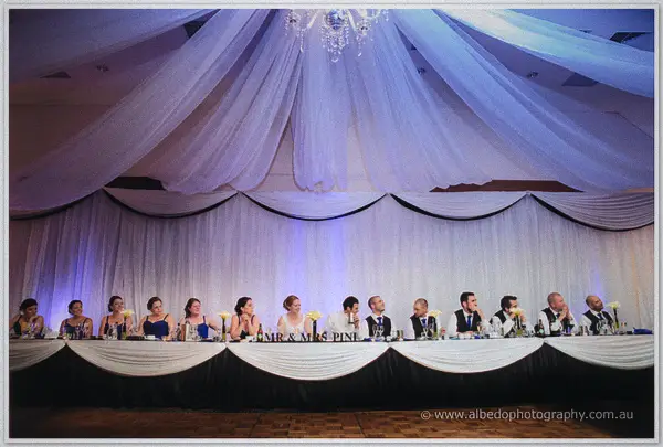 The long bridal table glows under a crystal chandelier and Mr and Mrs lettering as guests toast beneath drapes