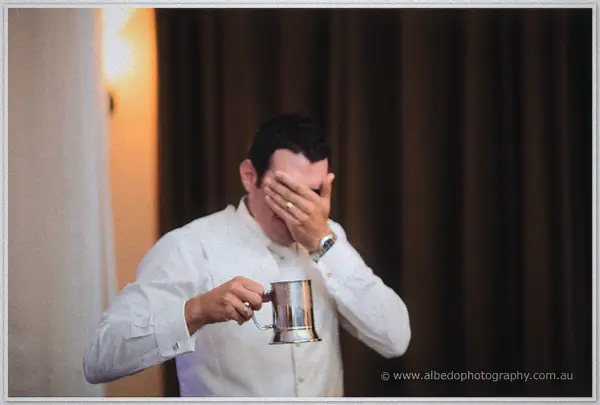 Joe laughs into his hand while raising a silver tankard during the toasts, lamplight warm on the curtains