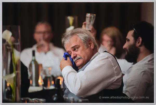 An older guest in a grey vest dabs his eyes during the speeches as friends toast behind him