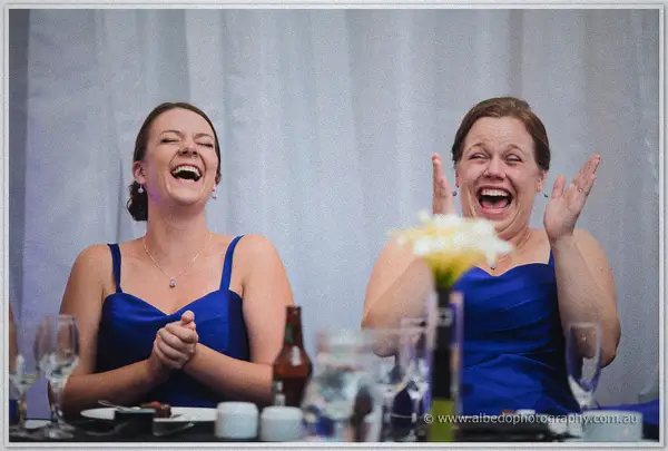 Two bridesmaids in royal blue laugh heartily at the bridal table, one clasping hands, one throwing arms high