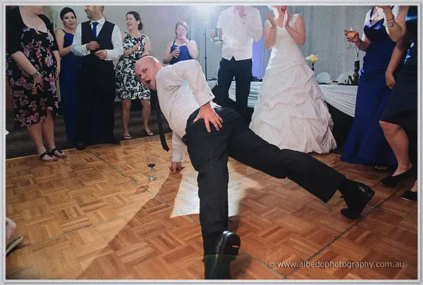 A bald groomsman strikes a playful low lunge on the parquet as cheering guests circle the dance floor
