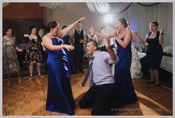 A groomsman kneels playfully between two bridesmaids in royal blue as guests dance and cheer around them