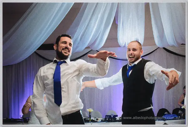 Two grinning groomsmen dance with arms outstretched in blue ties beneath the white ceiling drapes