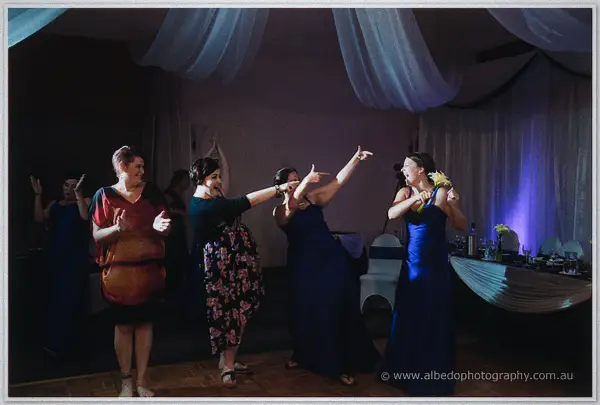 Dancing guests point toward the woman in blue clutching the caught bouquet under moody violet light